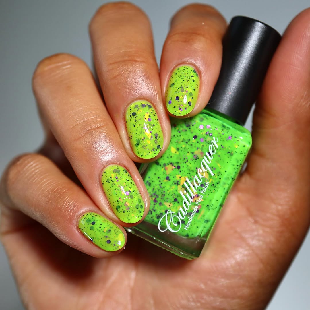Dapper but no Dan by Cadillacquer, neon lime green with dark and purple hex glitter particles and scattered yellow-orange iridescent flakes, bottle held in a light-medium hand