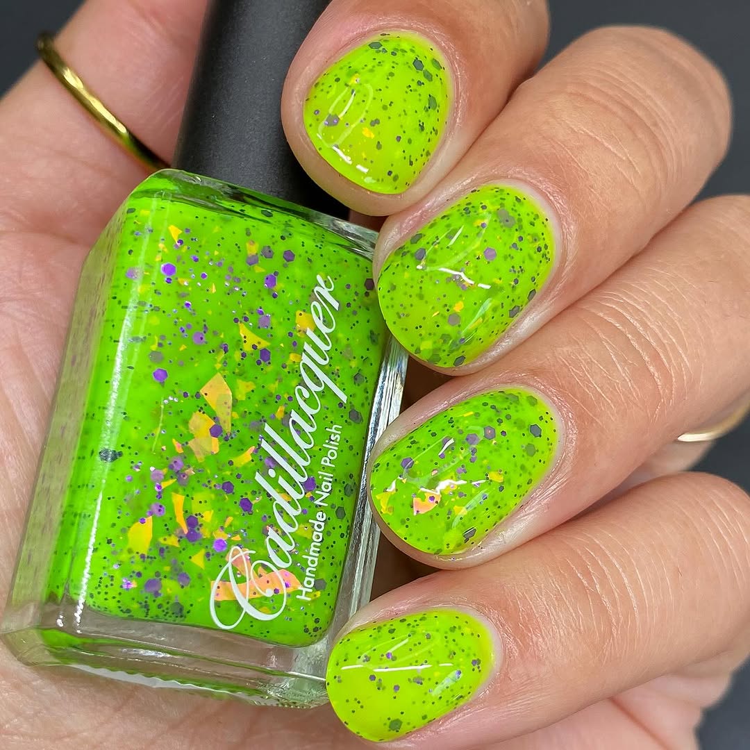 Dapper but no Dan by Cadillacquer, neon lime green jelly base with scattered purple hex glitter and large orange-yellow iridescent shards, bottle held in a light-medium hand