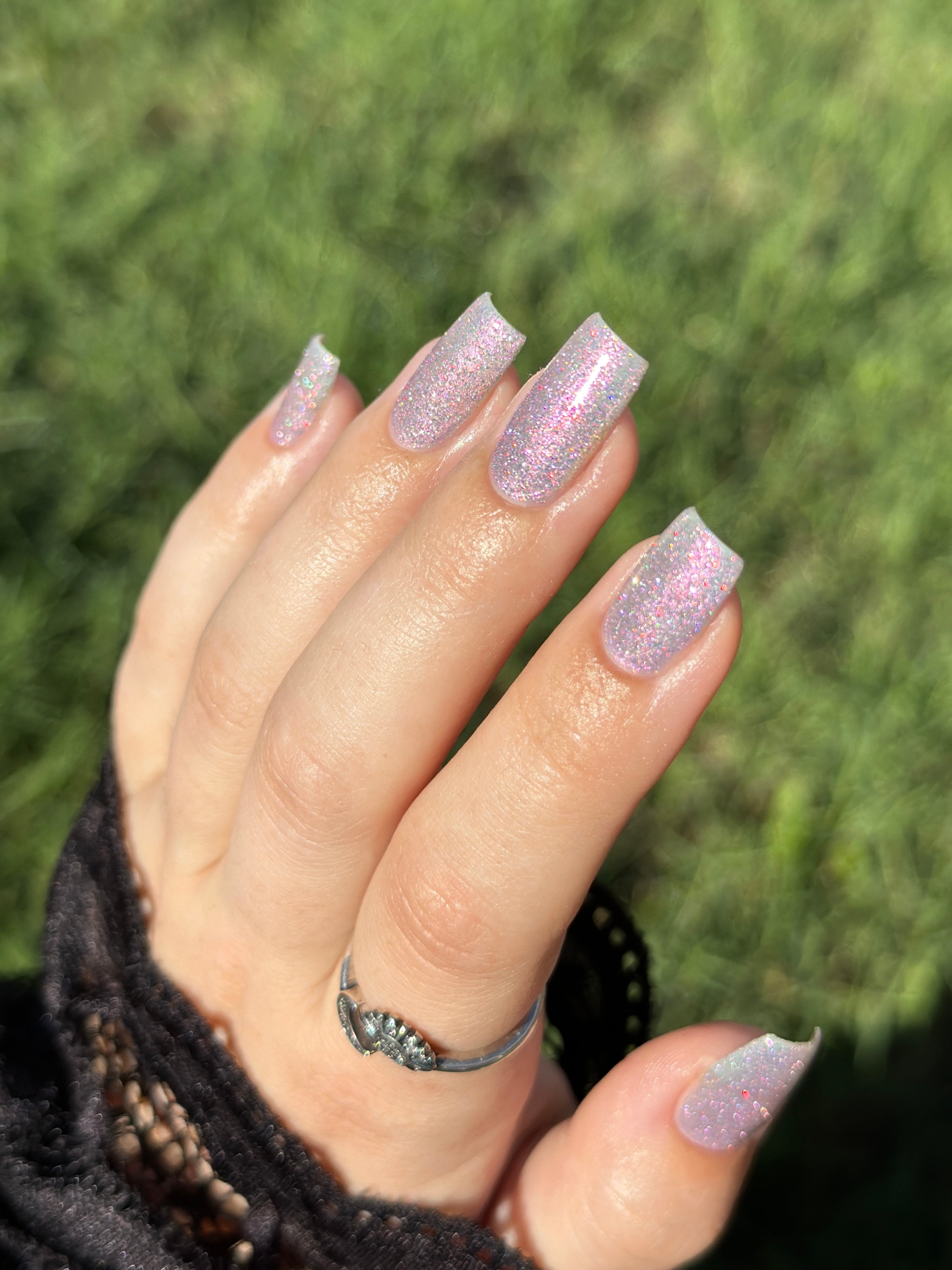 Lavender Haze by Glitch Lacquer on five square nails outdoors, very light purple polish with pink-yellow-green-teal multichrome reflect and red reflective glitters, light skin tone, bright daylight.