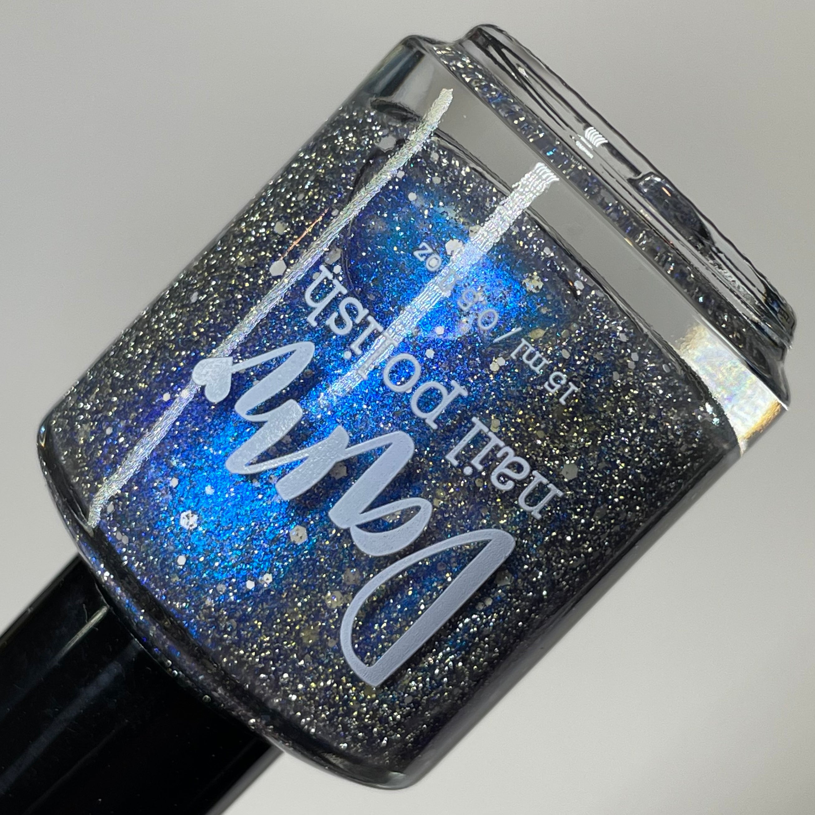 We're Off to See the Blizzard by Dam Nail Polish, deep black jelly base with blue-purple multichrome shimmer, silver reflective glitter, and scattered white glitter particles, bottle standalone product shot