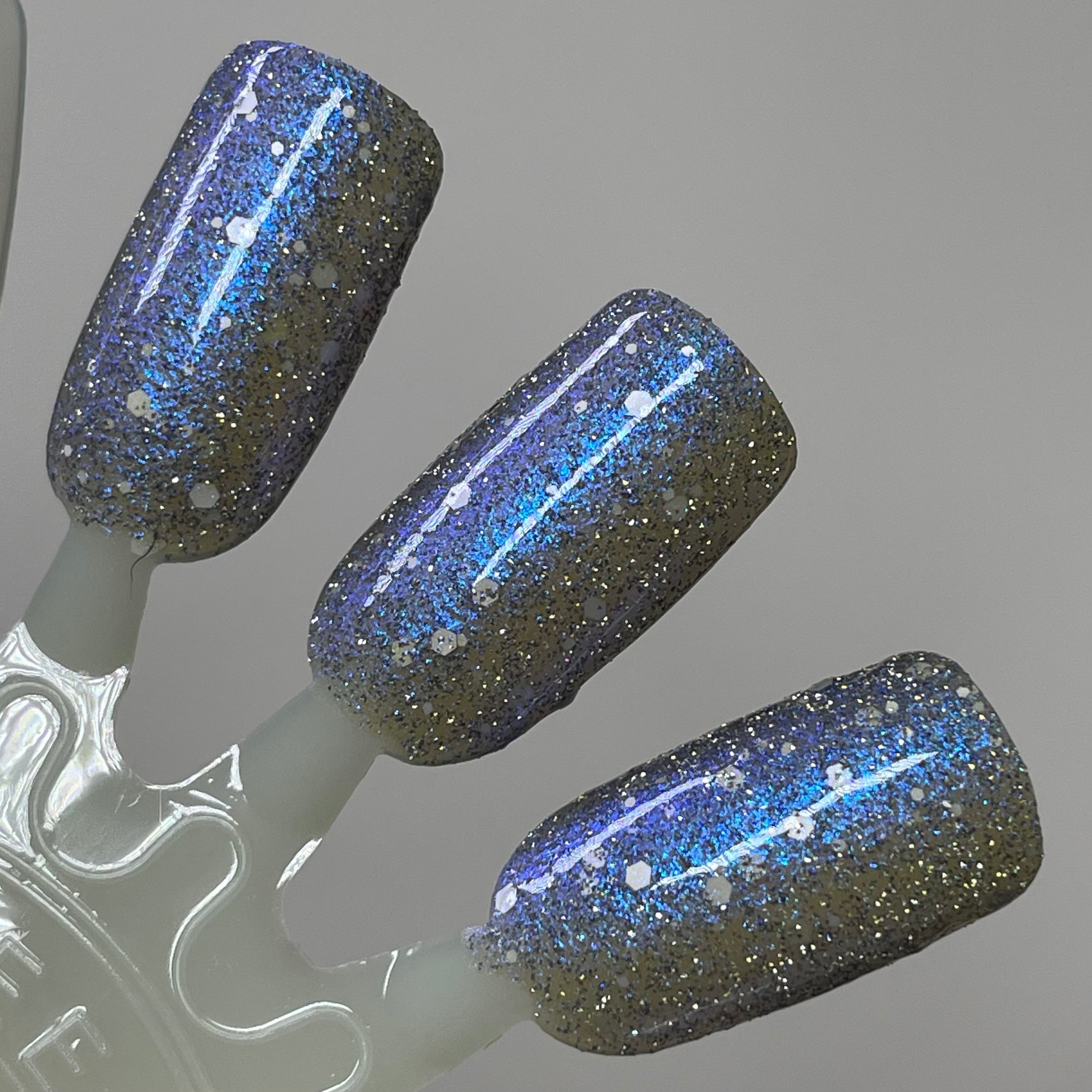 We're Off to See the Blizzard by Dam Nail Polish, deep charcoal-black jelly packed with silver micro-glitter, blue-violet multichrome shimmer streaks, and white hex glitters in studio light, swatch stick
