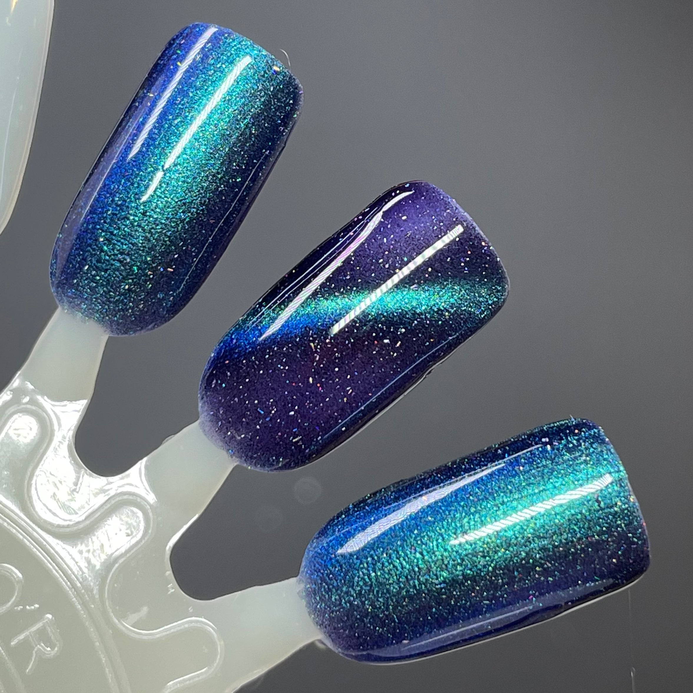 Wake Me Up Before You Cocoa by Dam Nail Polish, deep indigo-purple base with teal-to-blue linear magnetic pattern and holographic shimmer, swatch stick