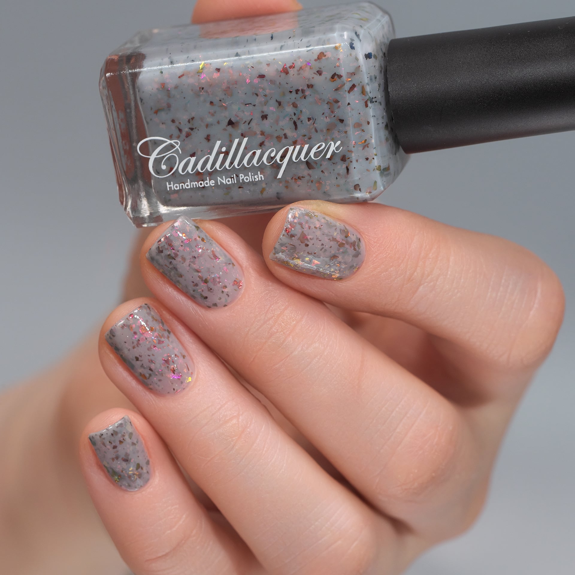 Cadillacquer Daydreaming grey red chameleon iridescent crelly nail polish, bottle held with four-nail swatch, light skin, studio lighting