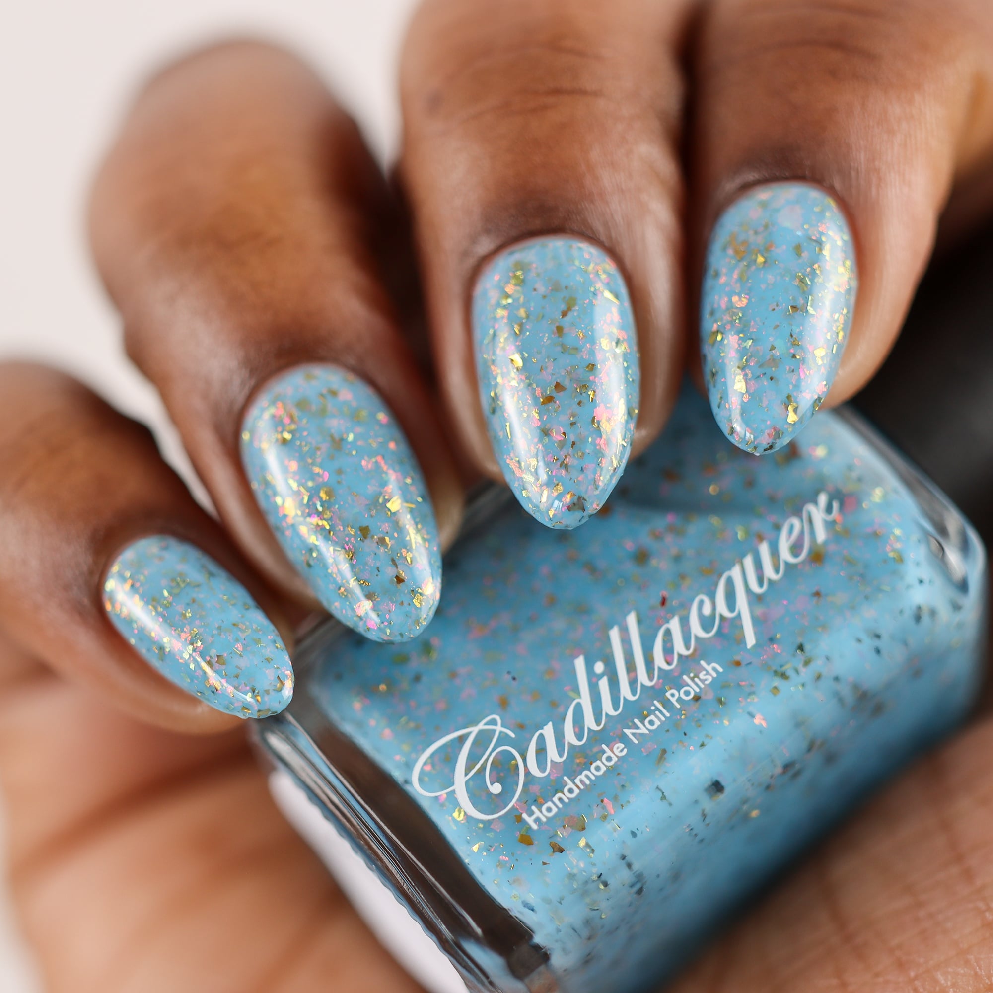 Cadillacquer Slow Mornings light blue gold chameleon iridescent crelly nail polish, bottle held with four-nail swatch, deep skin, studio lighting