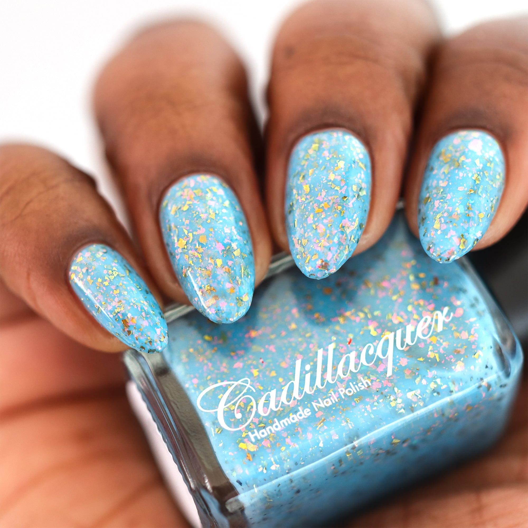 Cadillacquer Slow Mornings light blue gold chameleon iridescent crelly nail polish, bottle held with four-nail swatch, medium-deep skin, studio lighting