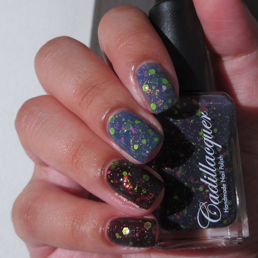 Thermal glitter and flake Cadillacquer nail polish in Strange Happenings, shown on four short natural nails with both warm (dusty blue) and cold (blackened eggplant) thermal states visible simultaneously, loaded with matte neon green hex glitter and pink iridescent flakes, with matching bottle held in hand, light-medium skin tone, indoor warm lighting, swatched by @_always_polished_
