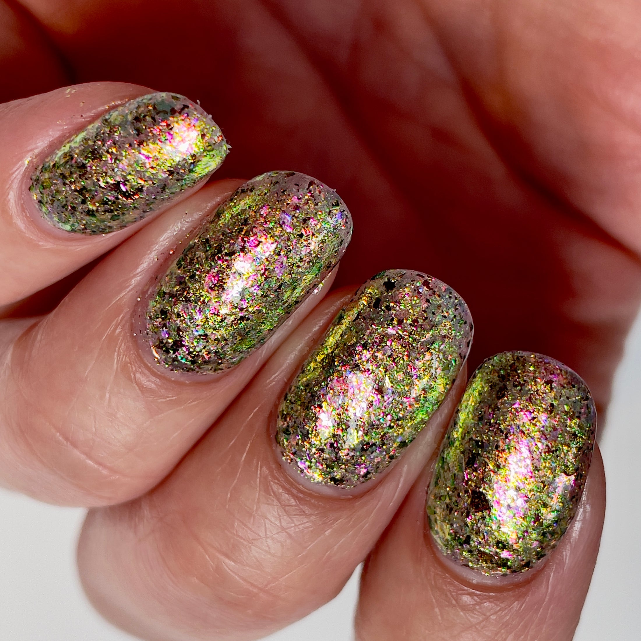 Ayla the Scoliosis Dolphin by Dam Nail Polish, multichrome flakie with gold, pink-magenta, and green-to-blue shifting flakies over a murky olive-khaki base under direct flash, on a light skin tone hand