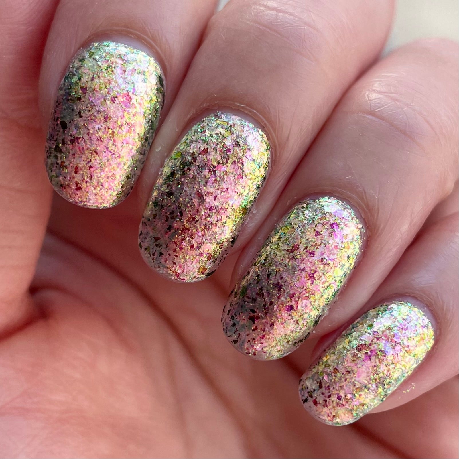 Ayla the Scoliosis Dolphin by Dam Nail Polish, multichrome flakie showing mixed-size pink, gold, and green-gold shifting flakies in bright daylight, on a light skin tone hand