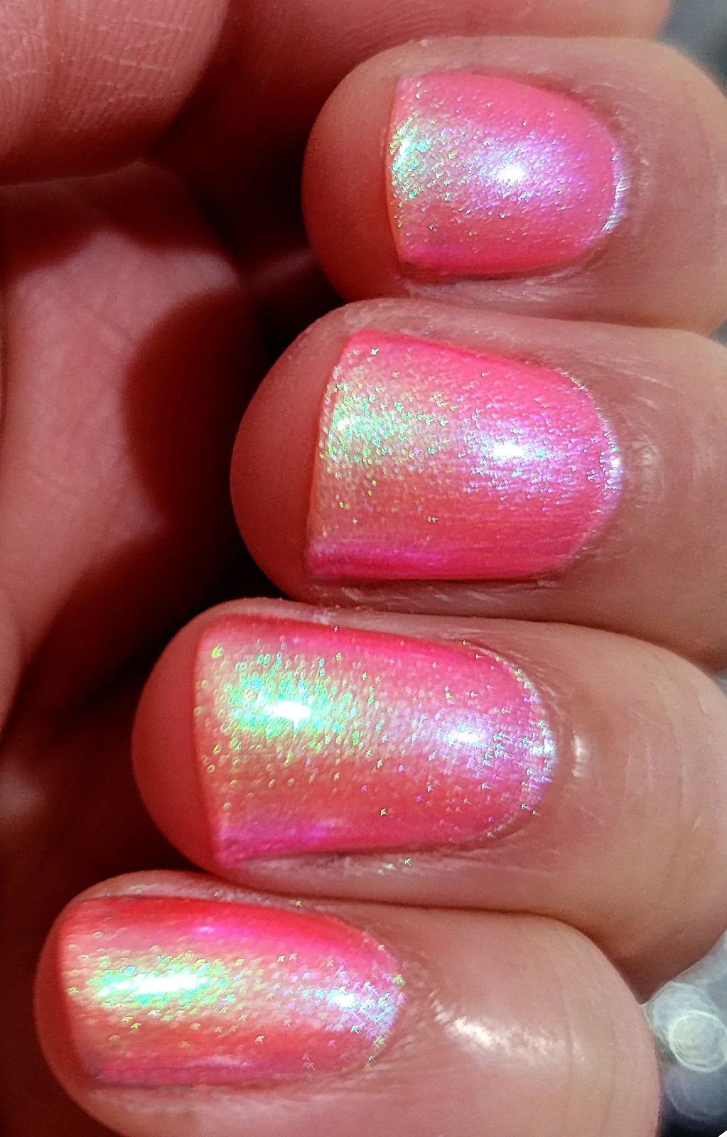 Nectar by Starrily, neon coral-pink base with vivid green-to-purple multichrome iridescent shimmer under indoor warm lighting, on a light-medium skin tone hand