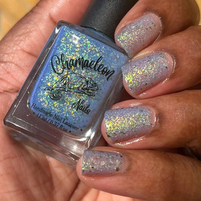 The Forgotten Lagoon (UV Polish) by Chamaeleon Nails, blue flakie UV in UV-activated state with vivid green-gold and orange iridescent flakes in bright daylight, bottle held in a medium-deep hand