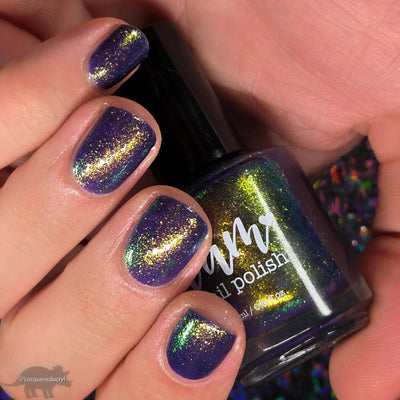 Sandman by Dam Nail Polish, deep blurple jelly with intense gold-to-green shifting shimmer, bottle held in a light skin tone hand