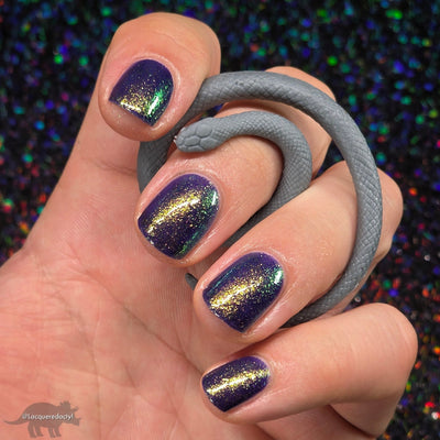 Sandman by Dam Nail Polish, deep blurple jelly with gold-to-green shifting shimmer streaks under direct flash, on a light skin tone hand