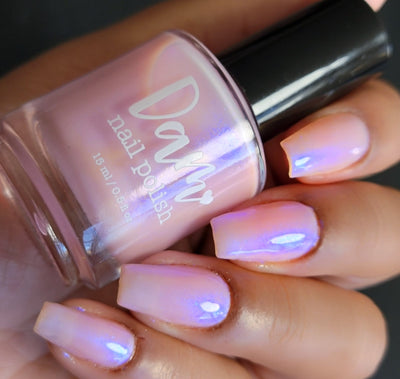 A Whimsical Dream by Dam Nail Polish, sheer blush pink shimmer with duochrome blurple flash amplified in bright daylight, bottle held in a light-medium skin tone hand