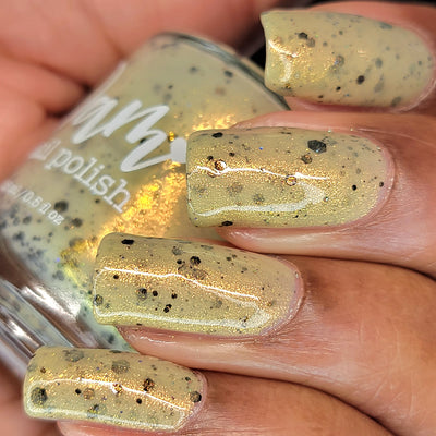 OG Devo-tea by Dam Nail Polish, tan-yellow crelly with gold shimmer and varied black glitter pieces showing crinkled flake detail in bottle, bottle held in a medium skin tone hand