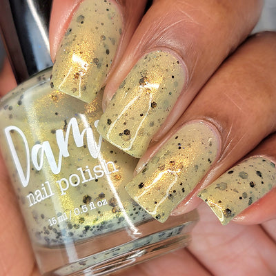 OG Devo-tea by Dam Nail Polish, pale yellow-tan crelly shimmer glitter with scattered black particles in daylight, bottle held in a medium skin tone hand