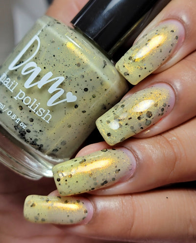 OG Devo-tea by Dam Nail Polish, pale yellow-green crelly with warm gold shimmer and scattered black hex and micro glitters, bottle held in a light-medium skin tone hand