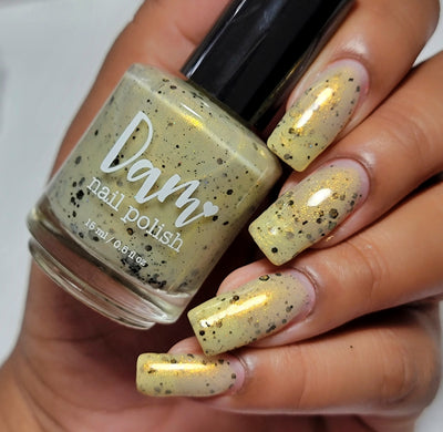 OG Devo-tea by Dam Nail Polish, pale yellow-tan crelly shimmer glitter with specular gold highlights under studio light, bottle held in a medium skin tone hand
