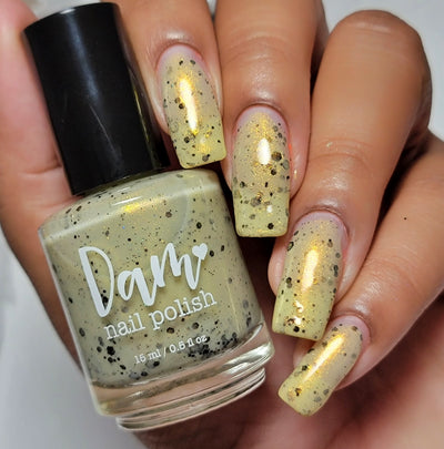 OG Devo-tea by Dam Nail Polish, tan crelly with gold shimmer and scattered black hex glitters, bottle held in a medium skin tone hand
