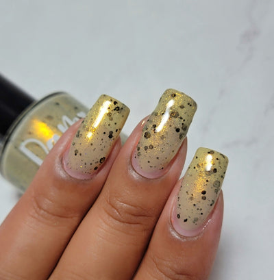 OG Devo-tea by Dam Nail Polish, warm tan-gold crelly with jelly-like translucency, gold shimmer, and assorted black glitters, on a light-medium skin tone hand