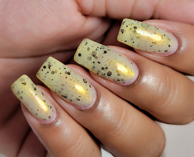 OG Devo-tea by Dam Nail Polish, pale yellow-green semi-translucent crelly with vivid gold shimmer flash and scattered black glitters in daylight, on a light-medium skin tone hand