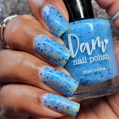 Blueberry Sanctuary by Dam Nail Polish, sheer light blue holographic shimmer crelly with mixed metallic blue hex and round glitters, bottle held in a medium-deep hand