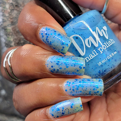 Blueberry Sanctuary by Dam Nail Polish, sheer light blue jelly crelly showing metallic blue hex glitters and holographic glitter dusting in bright daylight, bottle held in a medium hand