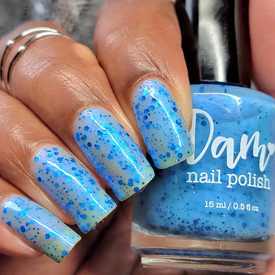 Blueberry Sanctuary by Dam Nail Polish, sheer light blue crelly with densely packed metallic blue hex glitters and blue-to-purple shifting shimmer, bottle held in a medium hand