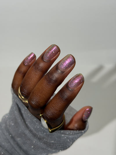 Lavender Haze by Glitch Lacquer on five squoval nails, very light purple polish with pink-yellow-green-teal multichrome reflect and red reflective glitters, deep skin tone, studio lighting.