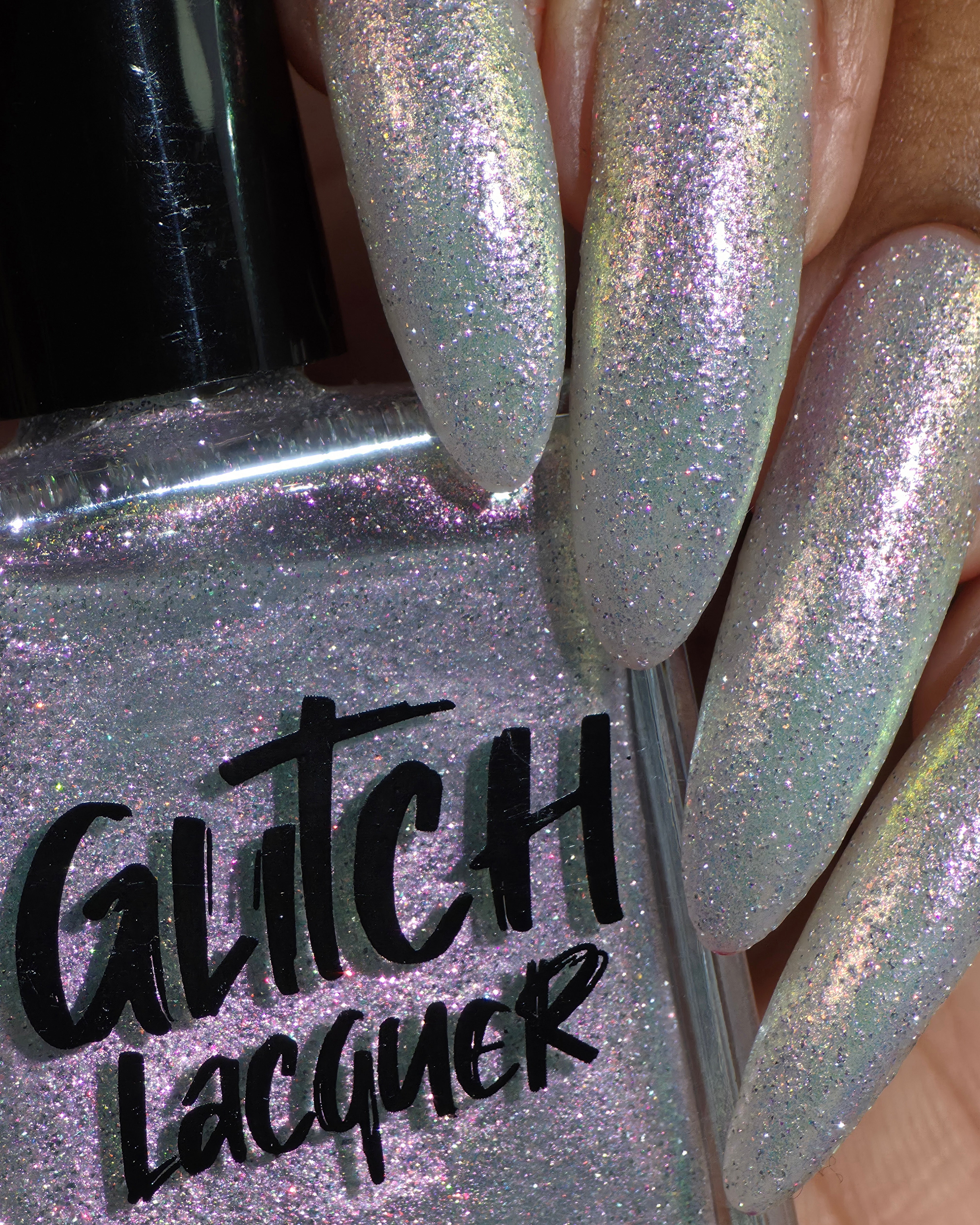 Lavender Haze by Glitch Lacquer on four long stiletto nails, very light purple polish with pink-yellow-green-teal multichrome reflect and red reflective glitters, medium-deep skin tone, studio lighting.