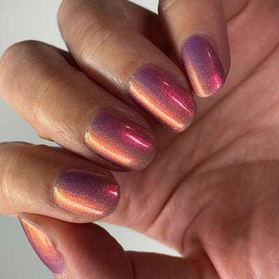 Maui by Dam Nail Polish, deep rose-pink multichrome shimmer shifting to warm gold-orange with sheer purple base, on a light-medium skin tone hand