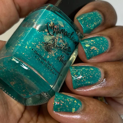 Kuang Si Falls by Chamaeleon Nails, neon teal jelly with rainbow iridescent flakies flashing golden-orange in bright daylight, bottle held in a medium-deep skin tone hand