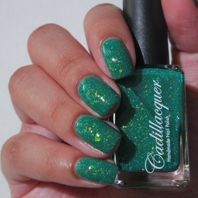 Blausee by Cadillacquer, teal green crelly with yellow-gold iridescent flakies and green reflective shimmer under indoor warm light, bottle held in a light hand