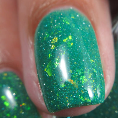Blausee by Cadillacquer, macro close-up of teal green crelly with large scattered yellow-green iridescent flakies and fine reflective glitter under indoor warm light