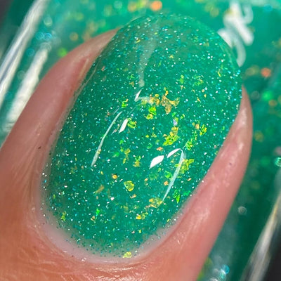 Blausee by Cadillacquer, teal green reflective glitter flakie crelly with light blue glitter flash and scattered yellow-green multichrome flakies under direct flash, on a light-medium skin tone hand
