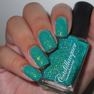 Blausee by Cadillacquer, teal green crelly showing scattered yellow-gold multichrome flakies and blue-green reflective shimmer under indoor warm light, bottle held in a light-medium hand