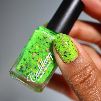 Dapper but no Dan by Cadillacquer, neon lime green with scattered black-purple glitter and orange-yellow iridescent flakes, bottle held in a medium-deep hand