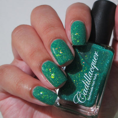 Blausee by Cadillacquer, teal green crelly showing yellow-green iridescent flakes and green reflective glitter under indoor warm light, bottle held in a light hand