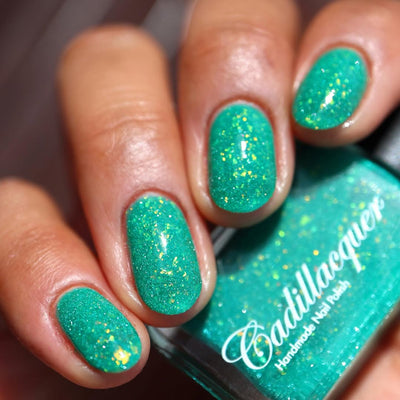 Blausee by Cadillacquer, teal green crelly showing yellow-green flake shift and teal reflective glitter under indoor warm light, on a light-medium skin tone hand