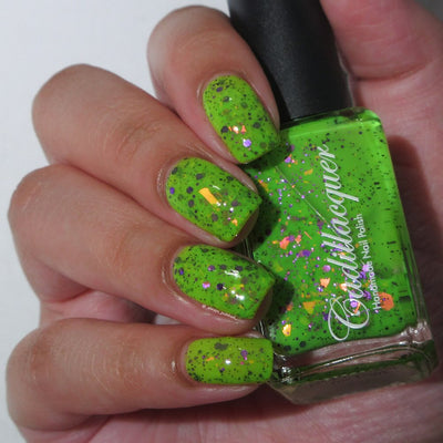Dapper but no Dan by Cadillacquer, neon lime green with purple glitter, orange-to-yellow iridescent flake shards, and black micro-specks under warm indoor light, bottle held in a light hand