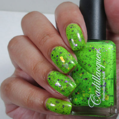 Dapper but no Dan by Cadillacquer, neon lime green with purple hex glitter, orange-yellow iridescent flakes, and small black scattered particles, bottle held in a light-medium hand