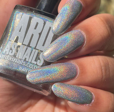 Cosmic by 'Ard As Nails, silver linear holographic with fine holographic particles and full-spectrum holo scatter in direct daylight, bottle held in a light-medium hand