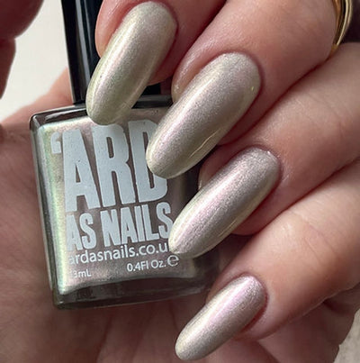Blushing Bridesmaid by 'Ard As Nails, muted champagne-beige base with soft iridescent pink-to-green shimmer topper in warm indoor light, bottle held in a light skin tone hand