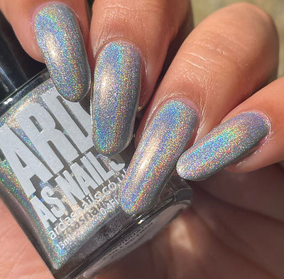 Cosmic by 'Ard As Nails, silver-grey linear holographic with vivid full-spectrum rainbow scatter in bright daylight, bottle held in a light-medium hand