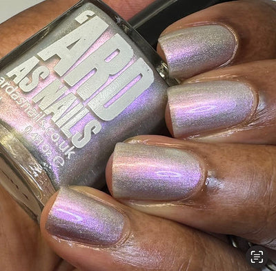 Blushing Bridesmaid by 'Ard As Nails, pink-to-green holographic duochrome shimmer topper showing lavender-pink shift under warm indoor light, bottle held in a deep skin tone hand