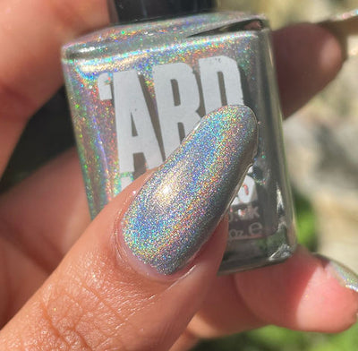 Cosmic by 'Ard As Nails, silver linear holographic with intense rainbow spectral scatter in bright daylight, bottle held in a medium hand