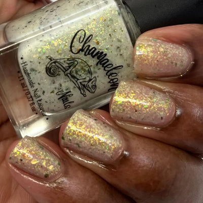 The Forgotten Lagoon (UV Polish) by Chamaeleon Nails, sheer off-white flakie UV base with gold, green, and orange-shifting ultrachrome flakes caught under direct flash, bottle held in a medium-deep hand