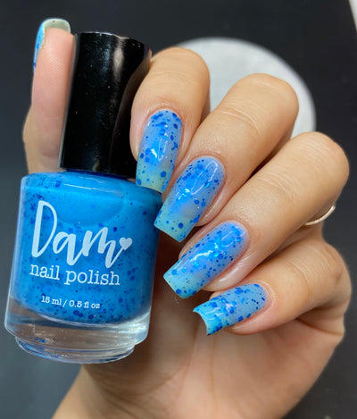 Blueberry Sanctuary by Dam Nail Polish, sheer blue crelly with metallic cobalt and holographic glitters showing pale icy shift near cuticle, bottle held in a light-medium hand