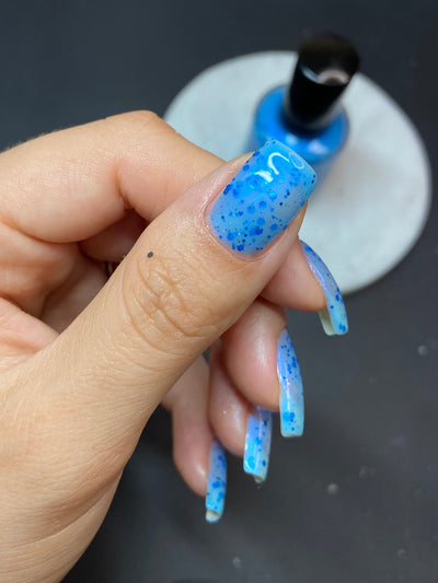 Blueberry Sanctuary by Dam Nail Polish, sheer light blue crelly with vivid cobalt metallic glitters and blue-to-purple color-shifting shimmer overlay, on a light-medium skin tone hand