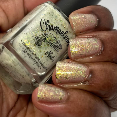 The Forgotten Lagoon (UV Polish) by Chamaeleon Nails, off-white flakie UV in resting state with green-to-orange ultrachrome flakes glowing warm gold and copper under indoor light, bottle held in a deep hand