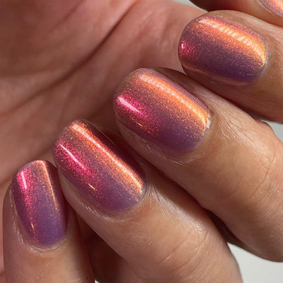 Maui by Dam Nail Polish, sheer purple-magenta multichrome shimmer shifting from hot pink to orange-gold, on a light-medium skin tone hand